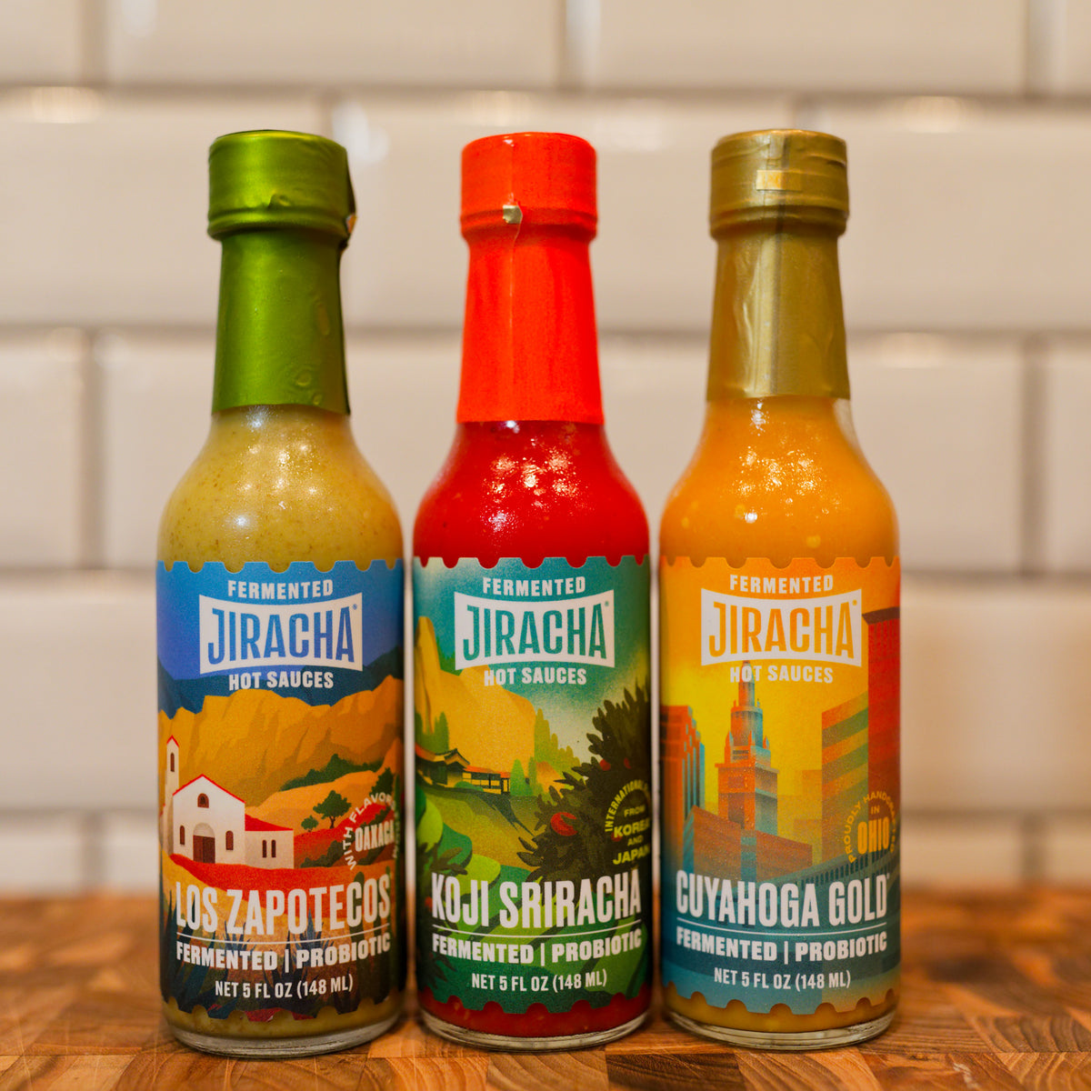 Three bottles of Jiracha hot sauces lined up on a wooden surface against a white subway tile wall: Los Zapotecos, Koji Sriracha, and Cuyahoga Gold.
