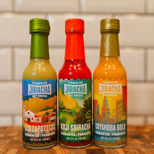 Three bottles of Jiracha hot sauces lined up on a wooden surface against a white subway tile wall: Los Zapotecos, Koji Sriracha, and Cuyahoga Gold.